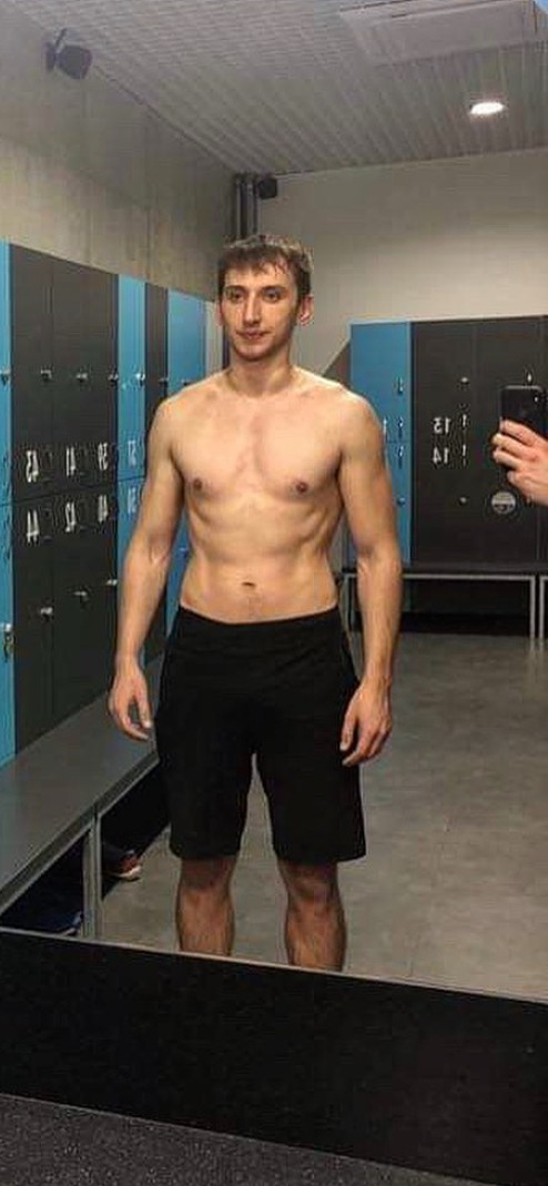 Mężczyzna w czarnych spodenkach prezentuje swoją sylwetkę w szatni z niebiesko-czarnymi szafkami, robiąc zdjęcie telefonem odbitym w lustrze.