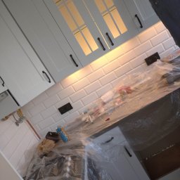 Damian Drankowski - Białe szafki kuchenne z podświetleniem, białe płytki ścienne typu metro, zabezpieczone folią malarską blaty i zlew w trakcie remontu.