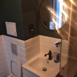 Nowoczesna łazienka z ciemnozieloną ścianą, białymi płytkami typu metro, okrągłym lustrem i umywalką z czarną armaturą, częściowo widoczna kabina prysznicowa.