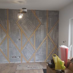 Damian Drankowski - Ściana w trakcie dekoracyjnego szpachlowania z geometrycznym wzorem wyznaczonym taśmą malarską, widoczne przygotowania i akcesoria malarskie.