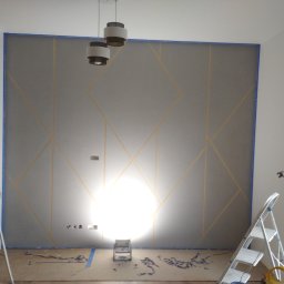 Damian Drankowski - Szara ściana z geometrycznym wzorem wykonanym za pomocą taśmy malarskiej, widoczne schodki malarskie i lampa halogenowa na podłodze, przygotowanie do malowania.