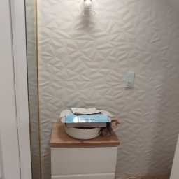 Damian Drankowski - Wnętrze łazienki w trakcie remontu: biała ściana z trójwymiarową fakturą, nowoczesna lampa ścienna, biała szafka z drewnianym blatem, na której leży umywalka i pudełko po baterii, widoczne kartony...