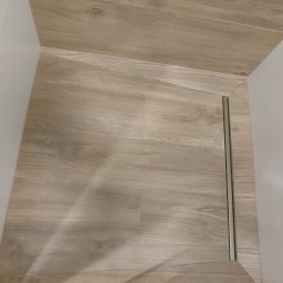 Damian Drankowski - Ukończona podłoga w łazience wyłożona panelami imitującymi drewno, widoczny odpływ liniowy przy kabinie prysznicowej oraz fragment buta osoby robiącej zdjęcie.