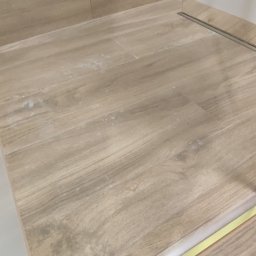 Damian Drankowski - Jasne płytki podłogowe imitujące drewno w łazience, z widocznym odpływem liniowym i fragmentem kabiny prysznicowej z elementami w kolorze złotym.