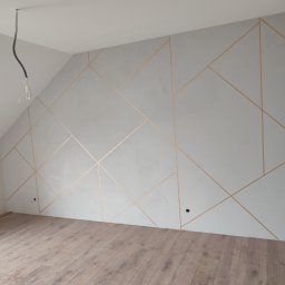Damian Drankowski - Ściana w pokoju na poddaszu z dekoracyjnym szpachlowaniem w geometryczne, złote linie; widoczne niedokończone gniazdka elektryczne i kabel zwisający z sufitu.