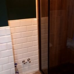 Damian Drankowski - Fragment łazienki z kabiną prysznicową z czarnymi ramami i białą, podłużną glazurą na ścianach, widoczne przyłącza wodne i fragment drewnopodobnej podłogi.