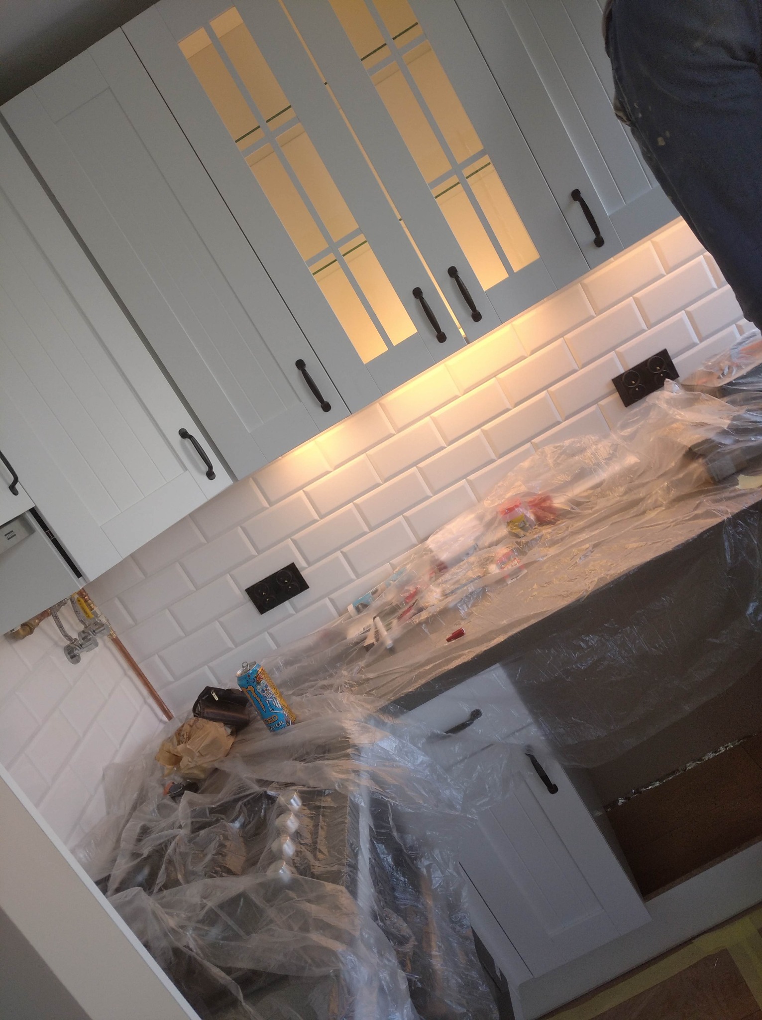 Białe szafki kuchenne z podświetleniem, białe płytki ścienne typu metro, zabezpieczone folią malarską blaty i zlew w trakcie remontu.