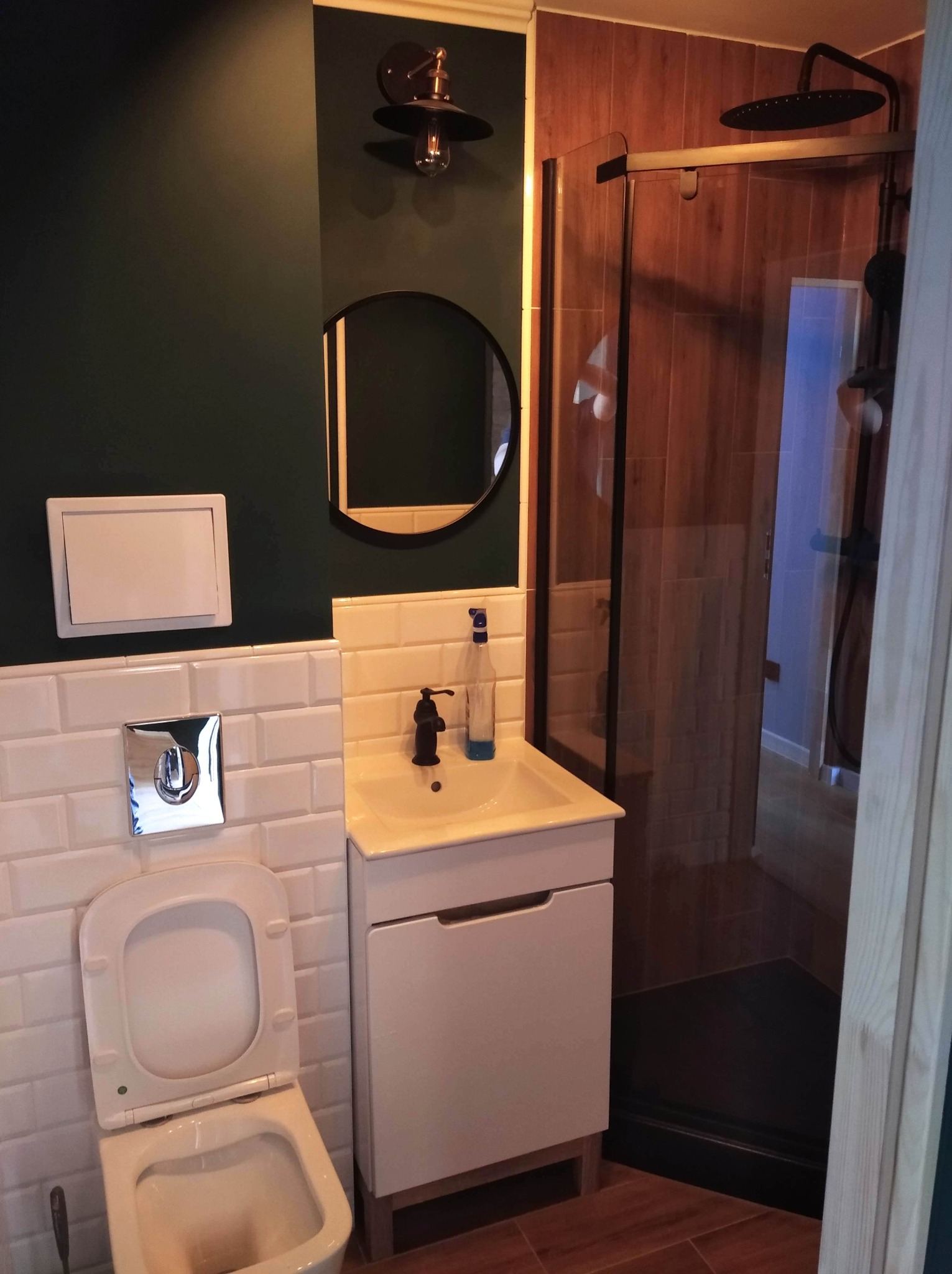 Nowoczesna aranżacja łazienki w Lubaniu: ciemnozielona ściana, białe płytki typu metro, okrągłe lustro, umywalka z szafką i kabina prysznicowa z drewnopodobnymi płytkami.