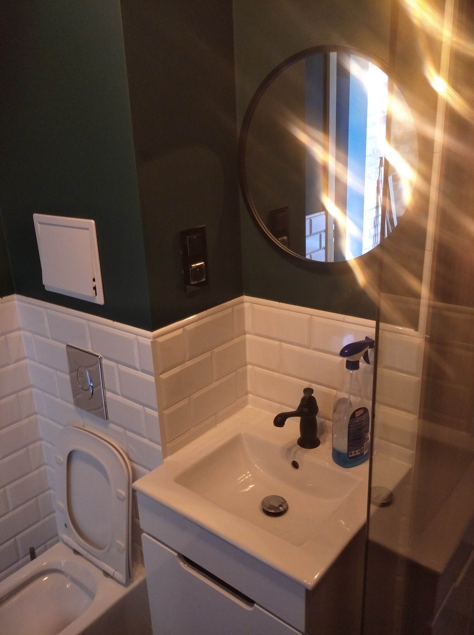 Nowoczesna łazienka z ciemnozieloną ścianą, białymi płytkami typu metro, okrągłym lustrem i umywalką z czarną armaturą, częściowo widoczna kabina prysznicowa.