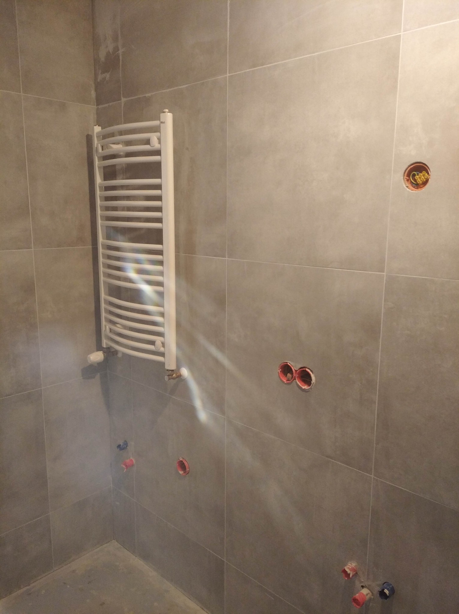 Szare płytki ścienne w łazience w trakcie remontu, widoczny grzejnik drabinkowy, puszki elektryczne i instalacja wodna przed montażem armatury.