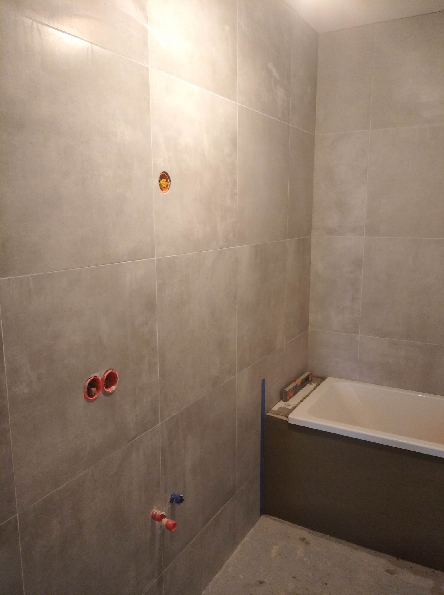 Łazienka w trakcie remontu: szare płytki ceramiczne na ścianach, widoczne otwory na instalacje elektryczne i wodne, częściowo obudowana wanna, poziomica przy wannie.