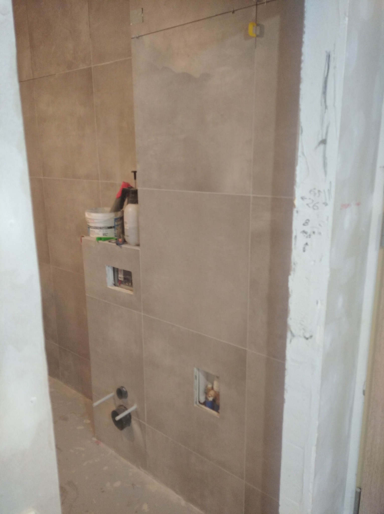 Wnętrze łazienki w trakcie remontu: ułożone płytki w odcieniach beżu, wystające rury instalacji hydraulicznej, pojemniki z materiałami budowlanymi na półce w zabudowie, ściana z widocznymi...