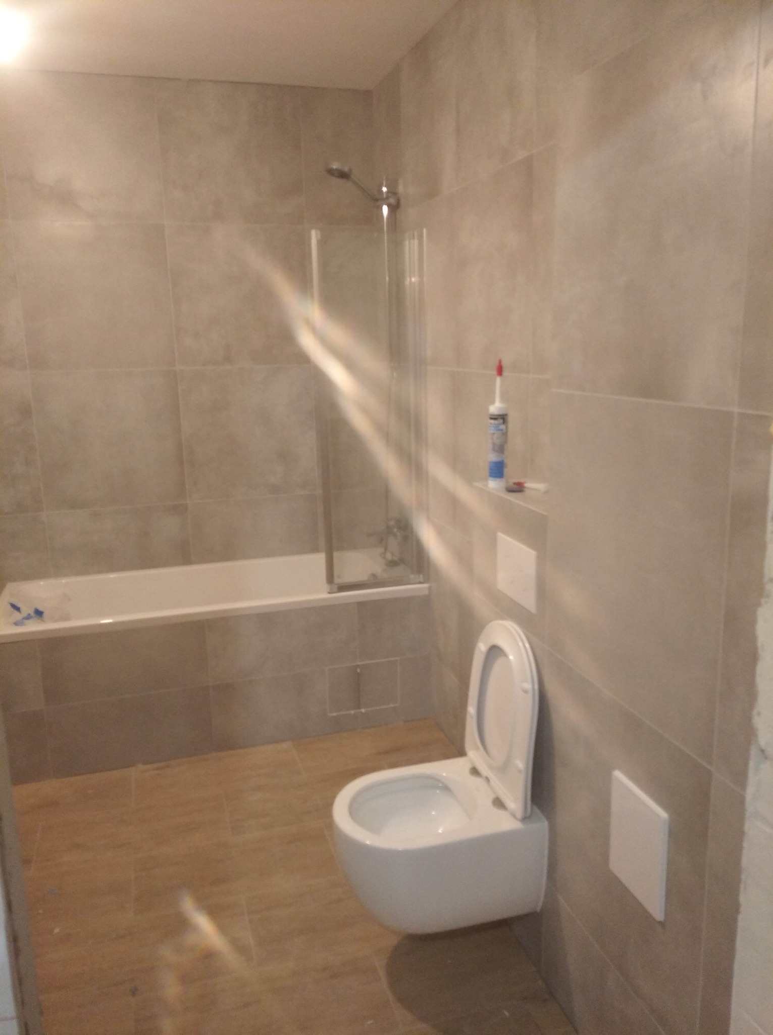 Wykończona łazienka w odcieniach beżu z podwieszaną toaletą, wanną z parawanem, i widoczną silikonową tubą na półce, ukazująca etap prac wykończeniowych.