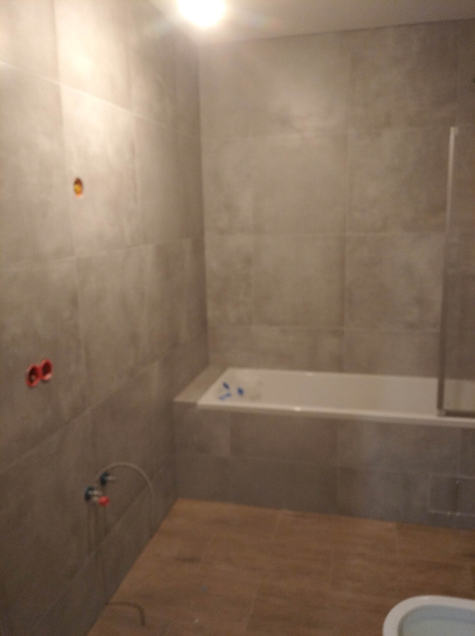 Wykończona łazienka z szarymi płytkami, widoczne przyłącza wodne, wanna z parawanem i fragment toalety w dolnym rogu kadru.