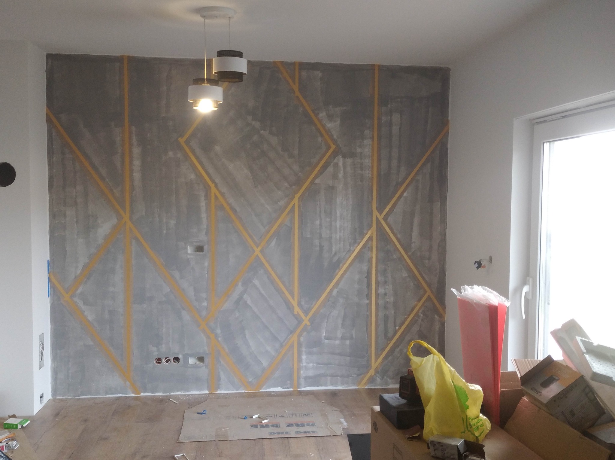 Ściana w trakcie dekoracyjnego szpachlowania z geometrycznym wzorem wyznaczonym taśmą malarską, widoczne przygotowania i akcesoria malarskie.