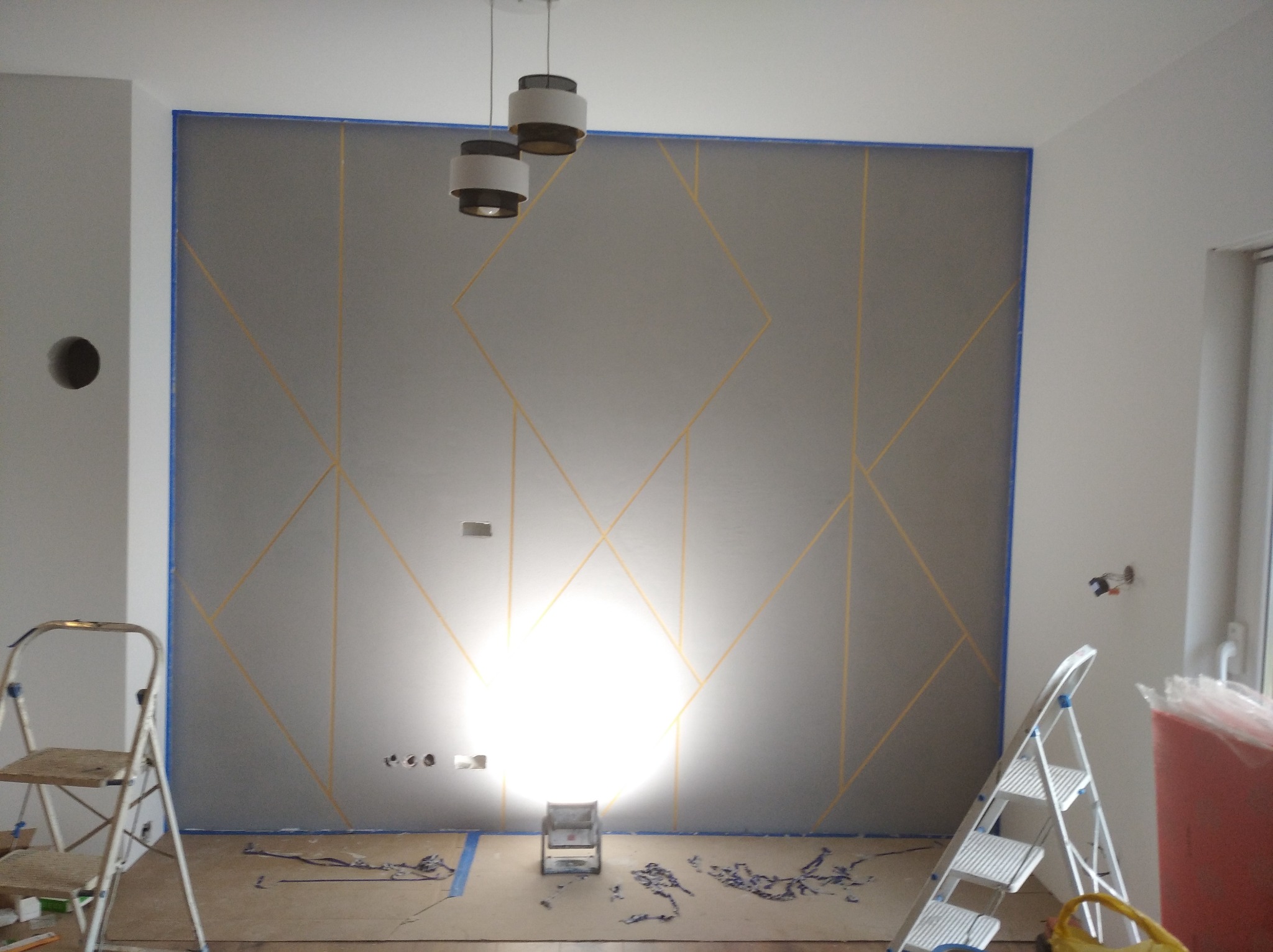 Szara ściana z geometrycznym wzorem wykonanym za pomocą taśmy malarskiej, widoczne schodki malarskie i lampa halogenowa na podłodze, przygotowanie do malowania.