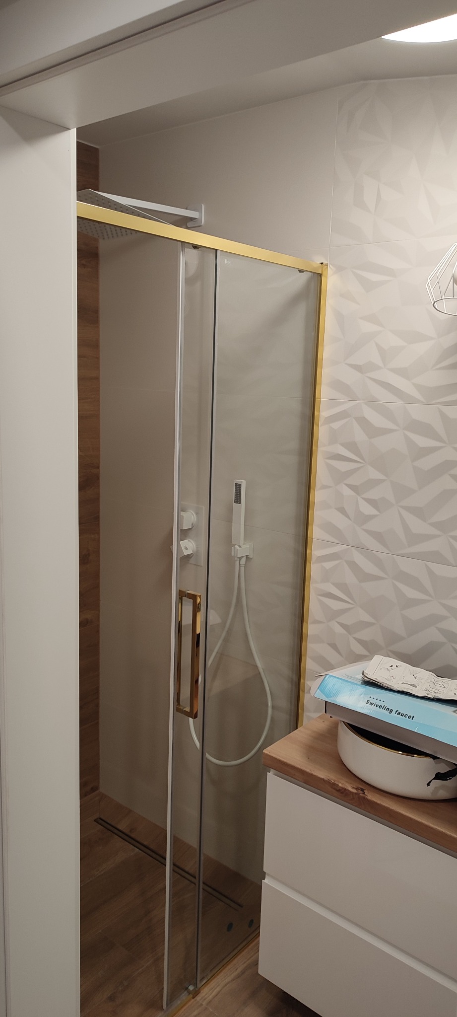 Nowoczesna kabina prysznicowa z złotymi profilami, widoczna armatura i odpływ liniowy w podłodze wyłożonej płytkami imitującymi drewno, na blacie pudełko z napisem 'Swiveling faucet'.