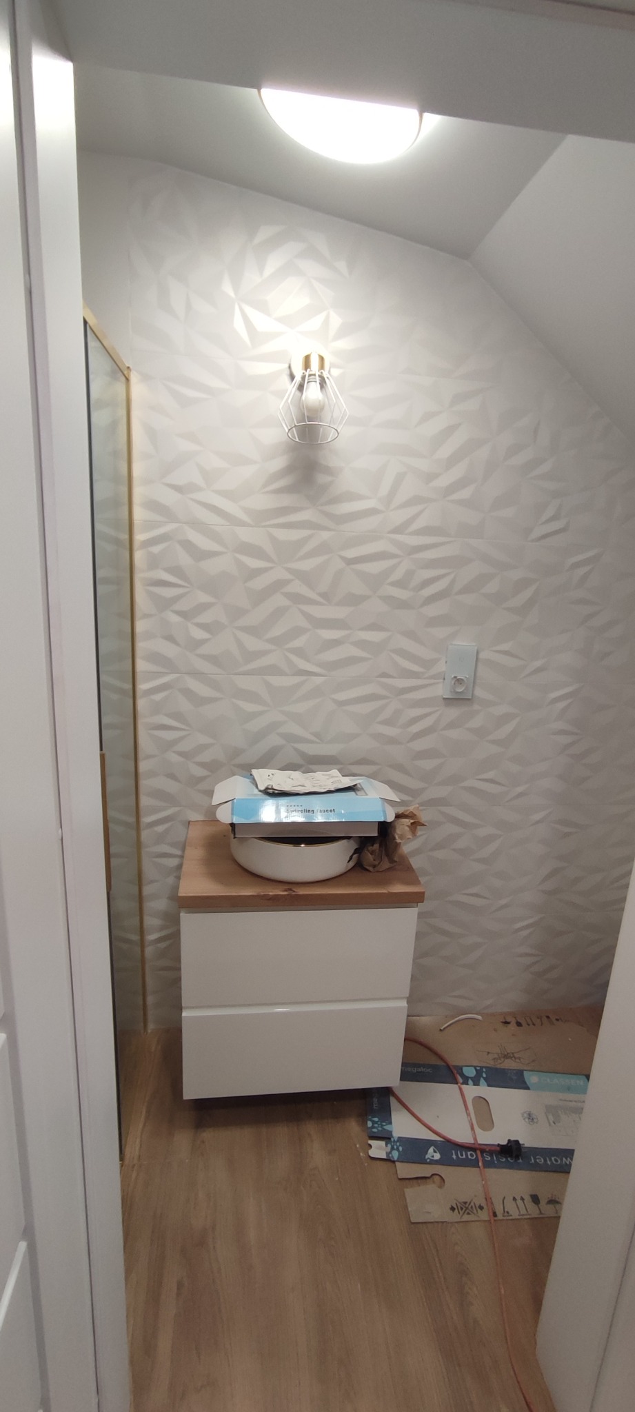Wnętrze łazienki w trakcie remontu: biała ściana z trójwymiarową fakturą, nowoczesna lampa ścienna, biała szafka z drewnianym blatem, na której leży umywalka i pudełko po baterii, widoczne kartony...