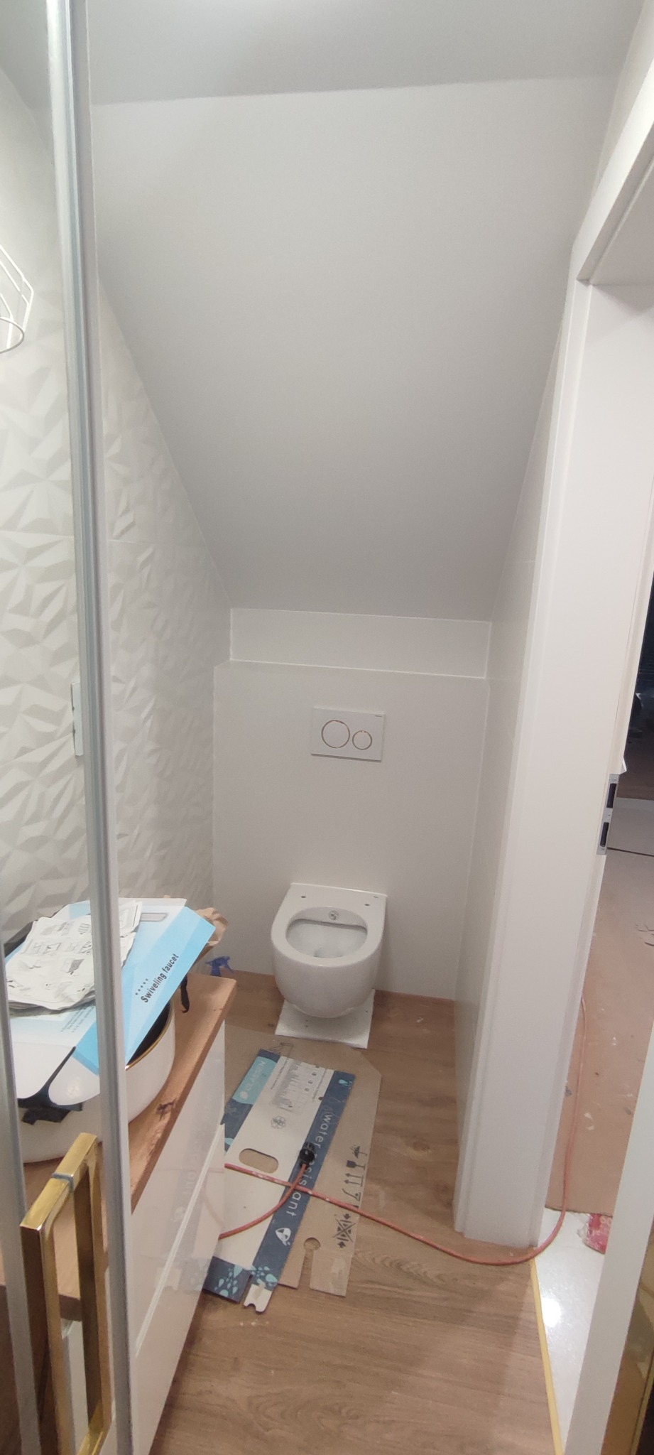 Nowa, biała toaleta bez deski zamontowana w małej łazience ze skośnym sufitem, widoczne kartony i narzędzia na podłodze.