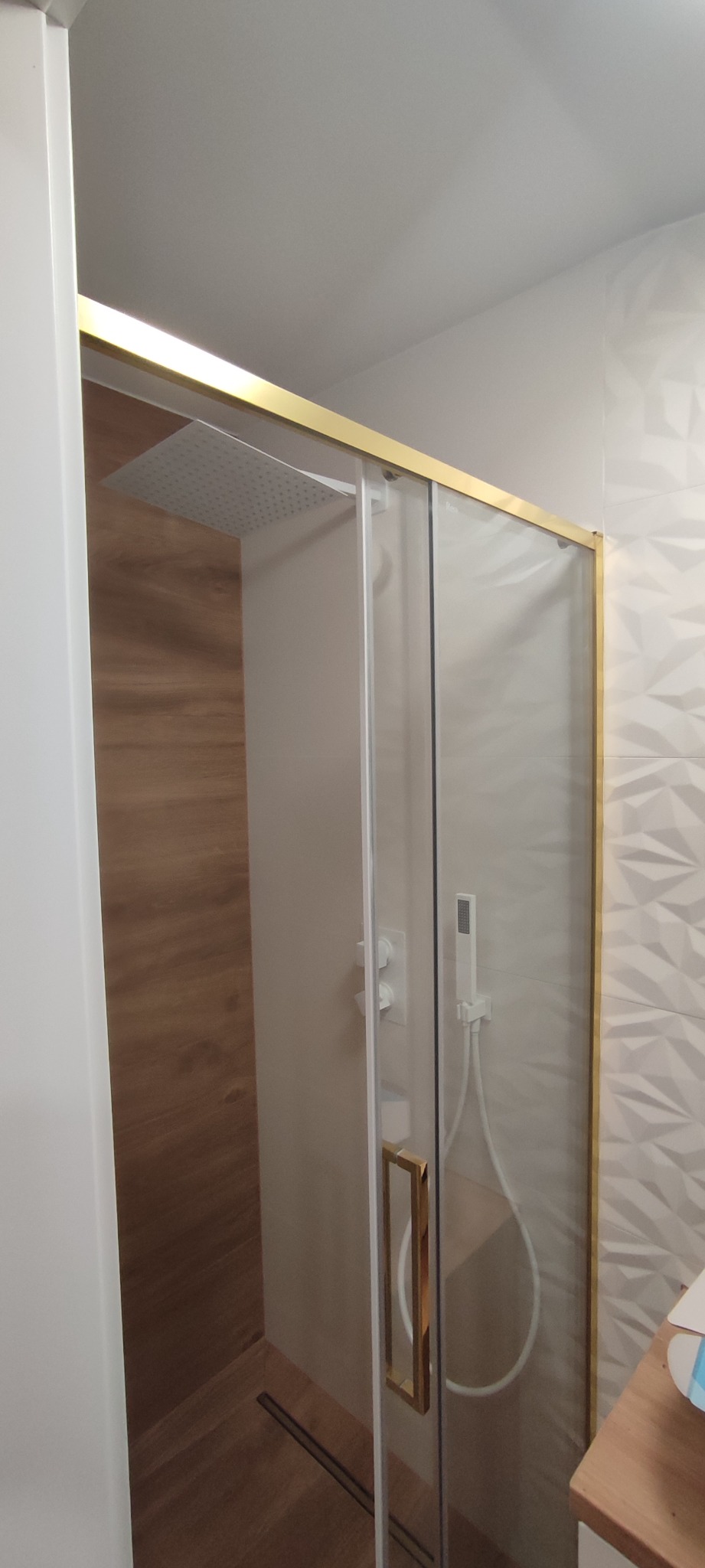 Nowoczesna kabina prysznicowa z przesuwnymi drzwiami w złotej ramie, deszczownicą i drewnopodobną ścianą, na tle białych płytek z geometrycznym wzorem.