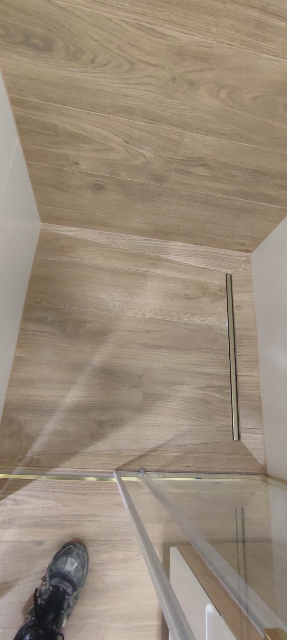 Ukończona podłoga w łazience wyłożona panelami imitującymi drewno, widoczny odpływ liniowy przy kabinie prysznicowej oraz fragment buta osoby robiącej zdjęcie.