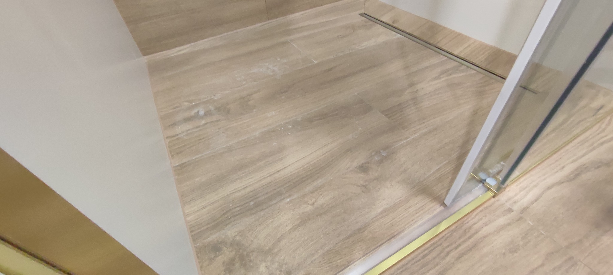 Jasne płytki podłogowe imitujące drewno w łazience, z widocznym odpływem liniowym i fragmentem kabiny prysznicowej z elementami w kolorze złotym.