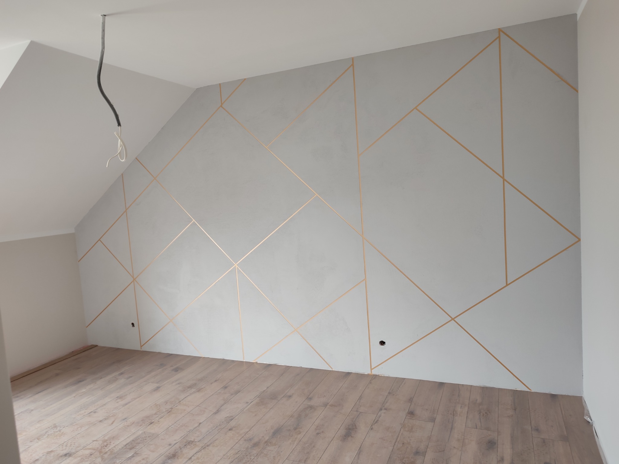 Ściana w pokoju na poddaszu z dekoracyjnym szpachlowaniem w geometryczne, złote linie; widoczne niedokończone gniazdka elektryczne i kabel zwisający z sufitu.