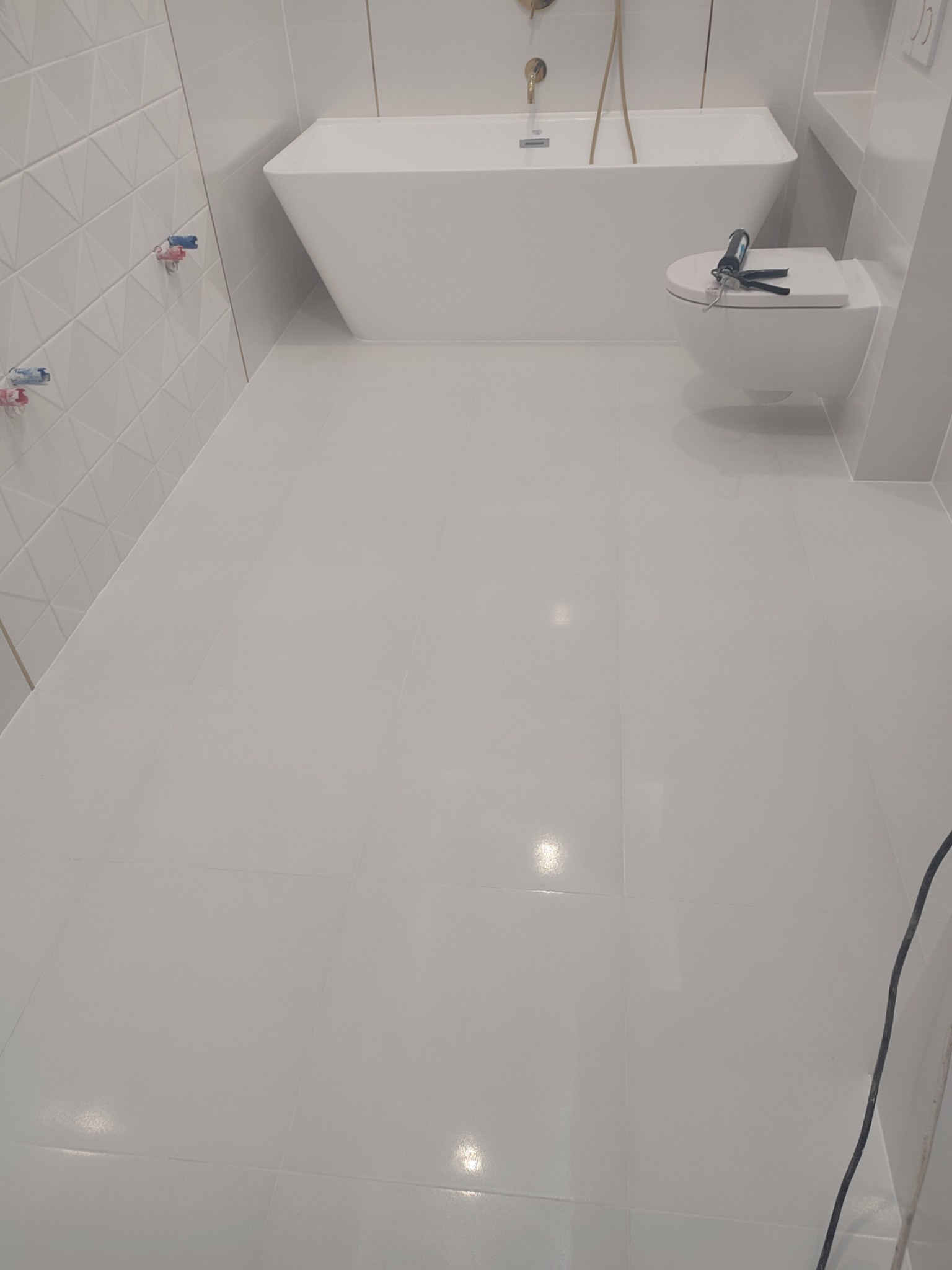 Biała, minimalistyczna łazienka po remoncie z błyszczącą podłogą z płytek, wiszącą muszlą klozetową z pistoletem do silikonu na desce, wolnostojącą wanną i geometryczną ścianą z płytek.