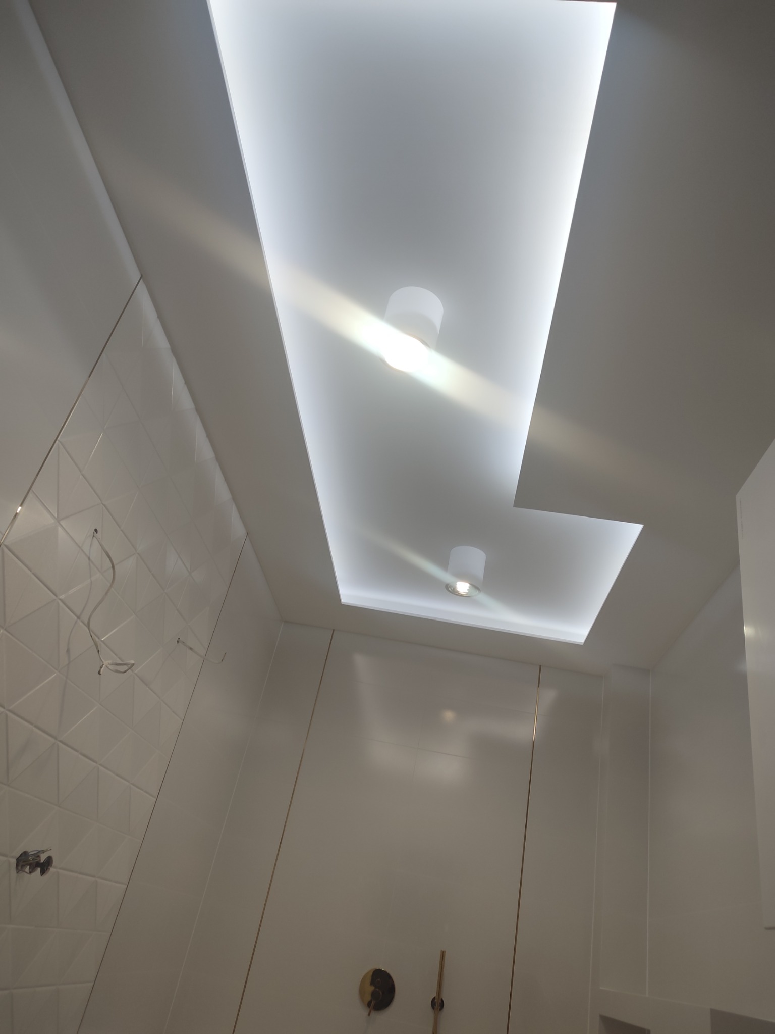 Nowoczesna łazienka z podwieszanym sufitem z oświetleniem LED i dwoma okrągłymi lampami punktowymi, białe płytki ścienne z geometrycznym wzorem, widoczne przewody instalacji elektrycznej