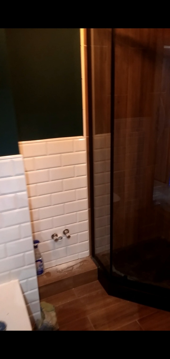 Fragment łazienki z kabiną prysznicową z czarnymi ramami i białą, podłużną glazurą na ścianach, widoczne przyłącza wodne i fragment drewnopodobnej podłogi.