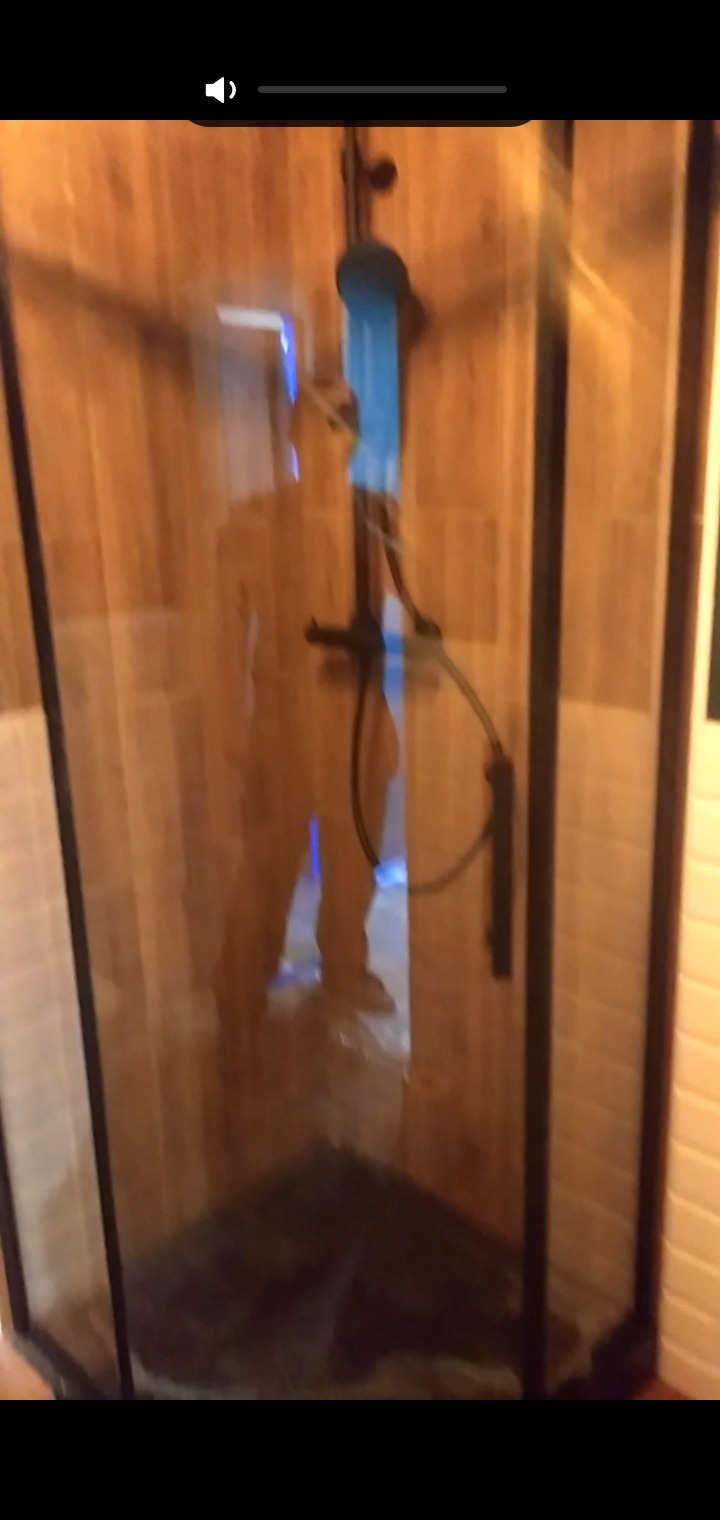 Kabina prysznicowa z czarnymi profilami i drewnianą ścianą w tle, widoczne elementy instalacji wodnej i osoba wewnątrz.