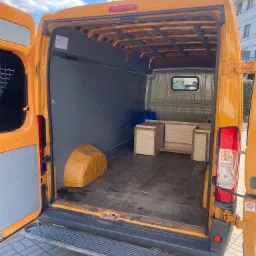 Wnętrze otwartego, pomarańczowego busa transportowego z drewnianymi elementami meblowymi w środku, zabezpieczonymi niebieską folią, widoczne z tyłu pojazdu na tle kostki brukowej i budynku.