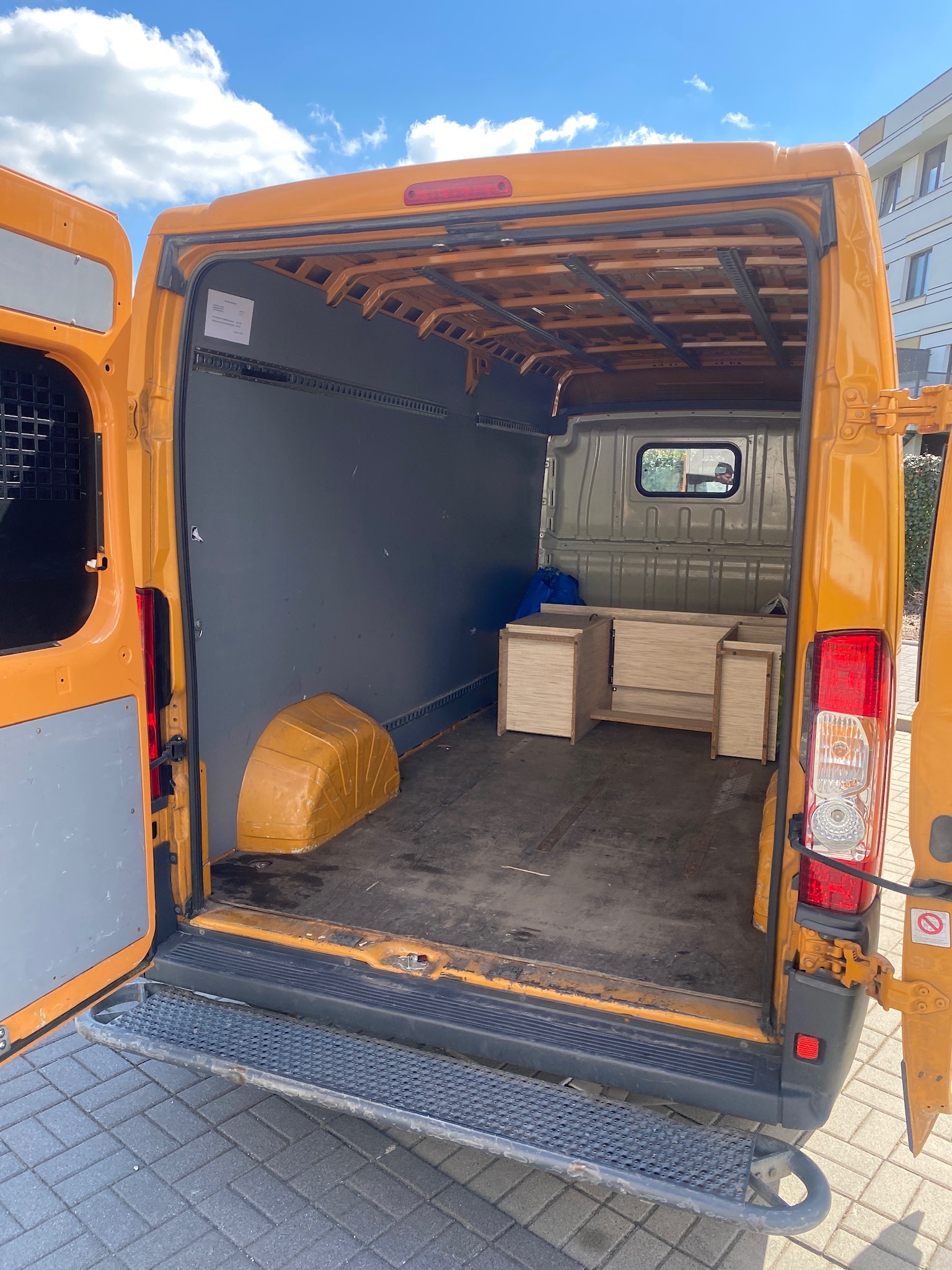 Wnętrze otwartego, pomarańczowego busa transportowego z drewnianymi elementami meblowymi w środku, zabezpieczonymi niebieską folią, widoczne z tyłu pojazdu na tle kostki brukowej i budynku.
