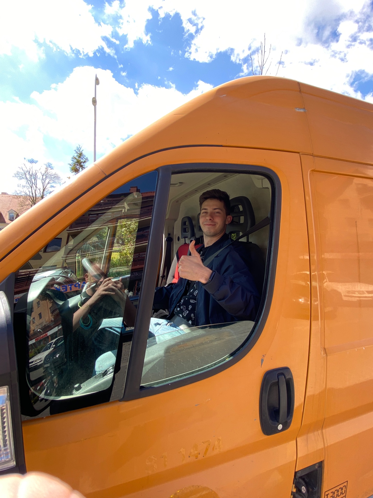 Uśmiechnięty kierowca w pomarańczowym busie z uniesionym kciukiem, widok z zewnątrz przez szybę, w tle błękitne niebo z chmurami.