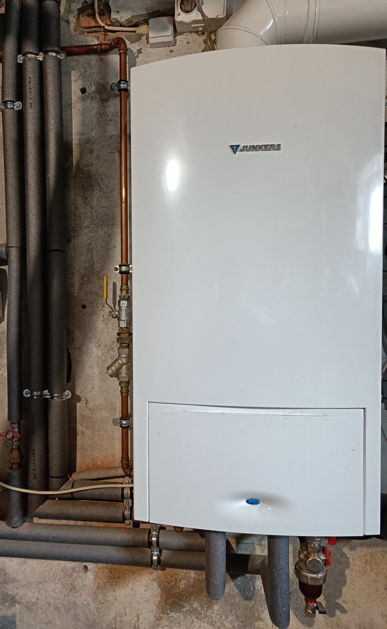 Biały piec gazowy marki Junkers zamontowany na surowej, betonowej ścianie, z widocznymi rurami miedzianymi i izolowanymi rurami grzewczymi.