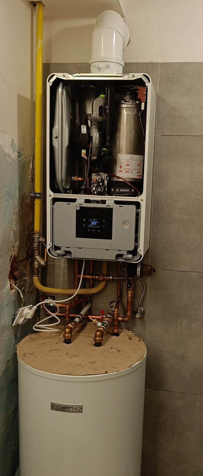 Otwarty, wiszący piec CO marki Bosch nad zasobnikiem ciepłej wody, z widocznymi rurami miedzianymi i żółtą rurą gazową, zamontowany w pomieszczeniu z szarą płytką na ścianie i fragmentem starej...