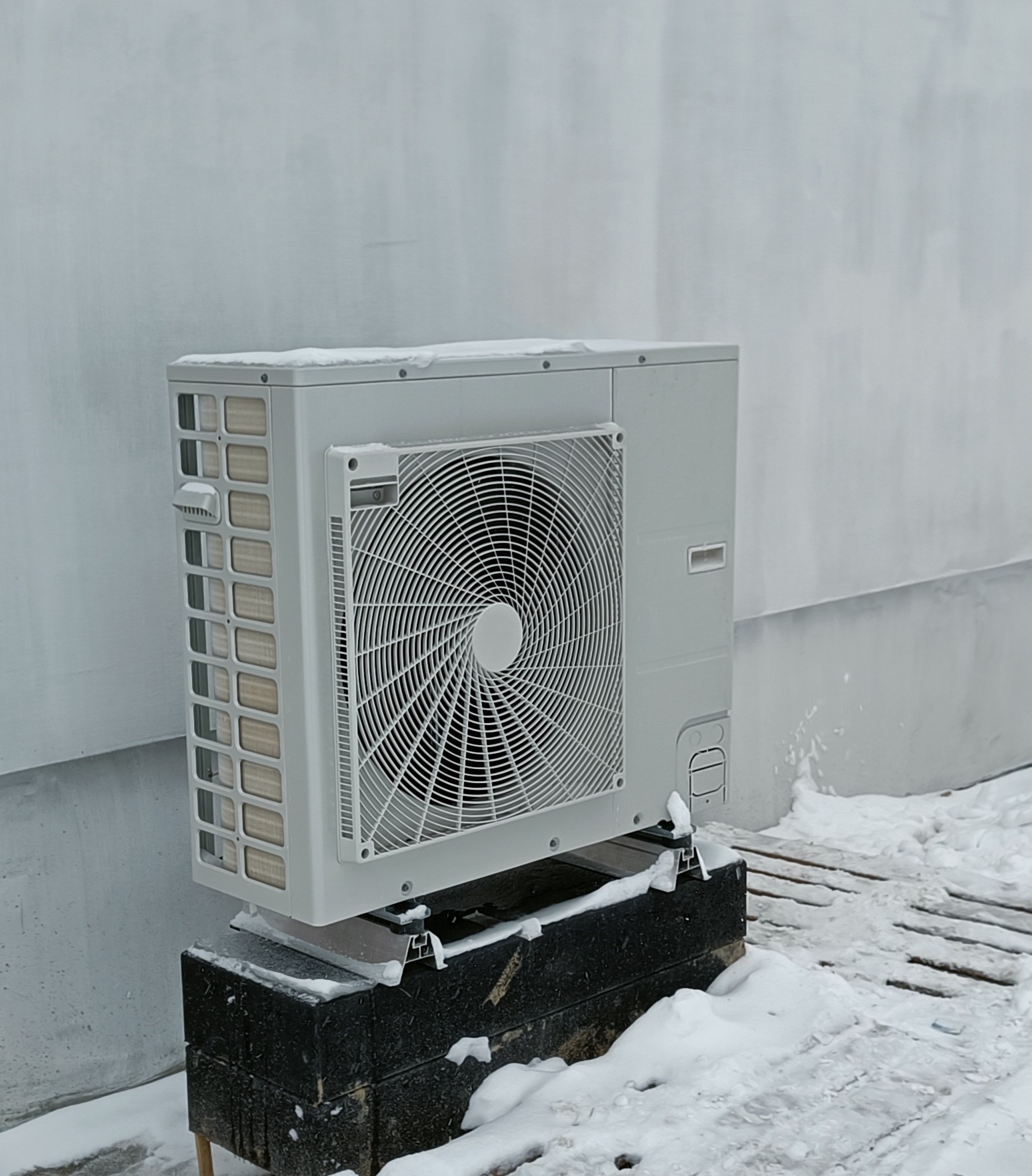Zewnętrzna jednostka klimatyzacji do domu, zamontowana na czarnym podwyższeniu, lekko przysypana śniegiem, na tle jasnej ściany budynku.