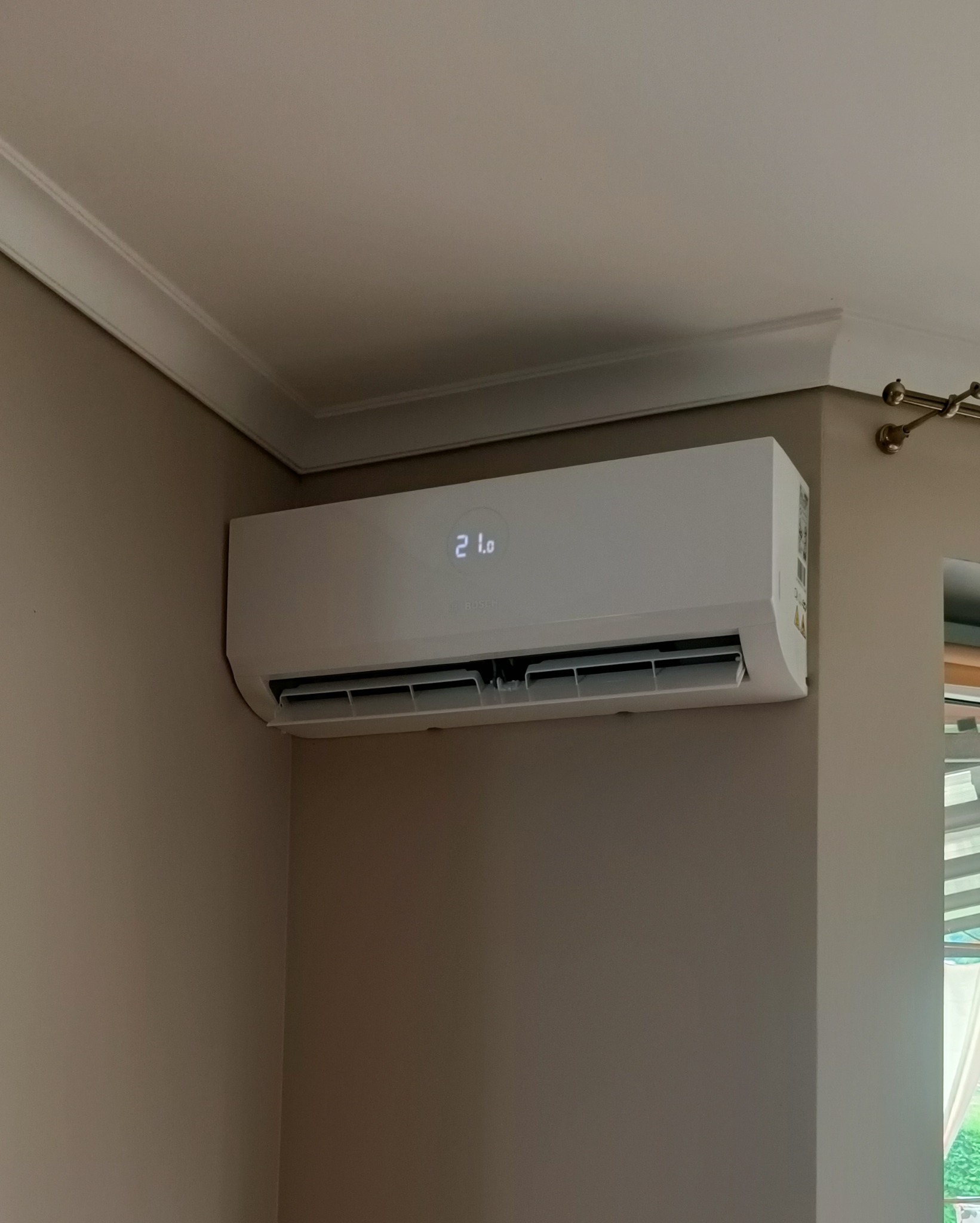 Biały klimatyzator ścienny marki Bosch zamontowany w rogu pokoju, wyświetlający temperaturę 21 stopni Celsjusza, widoczne listwy sufitowe i fragment okna.