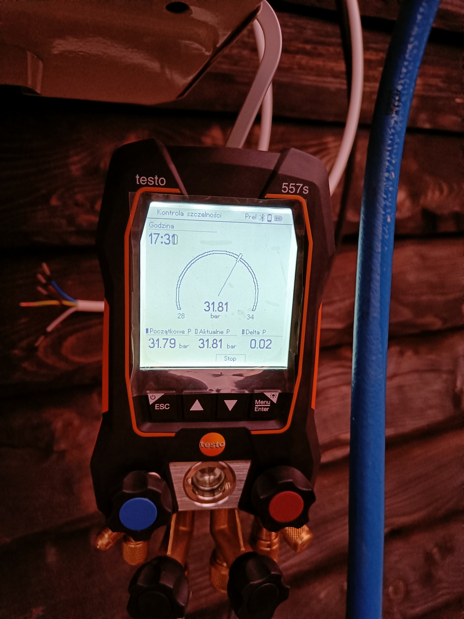 Cyfrowy manometr Testo 557s z wyświetlonymi wartościami ciśnienia podczas kontroli szczelności instalacji, widoczne przewody i fragment drewnianej ściany w tle.