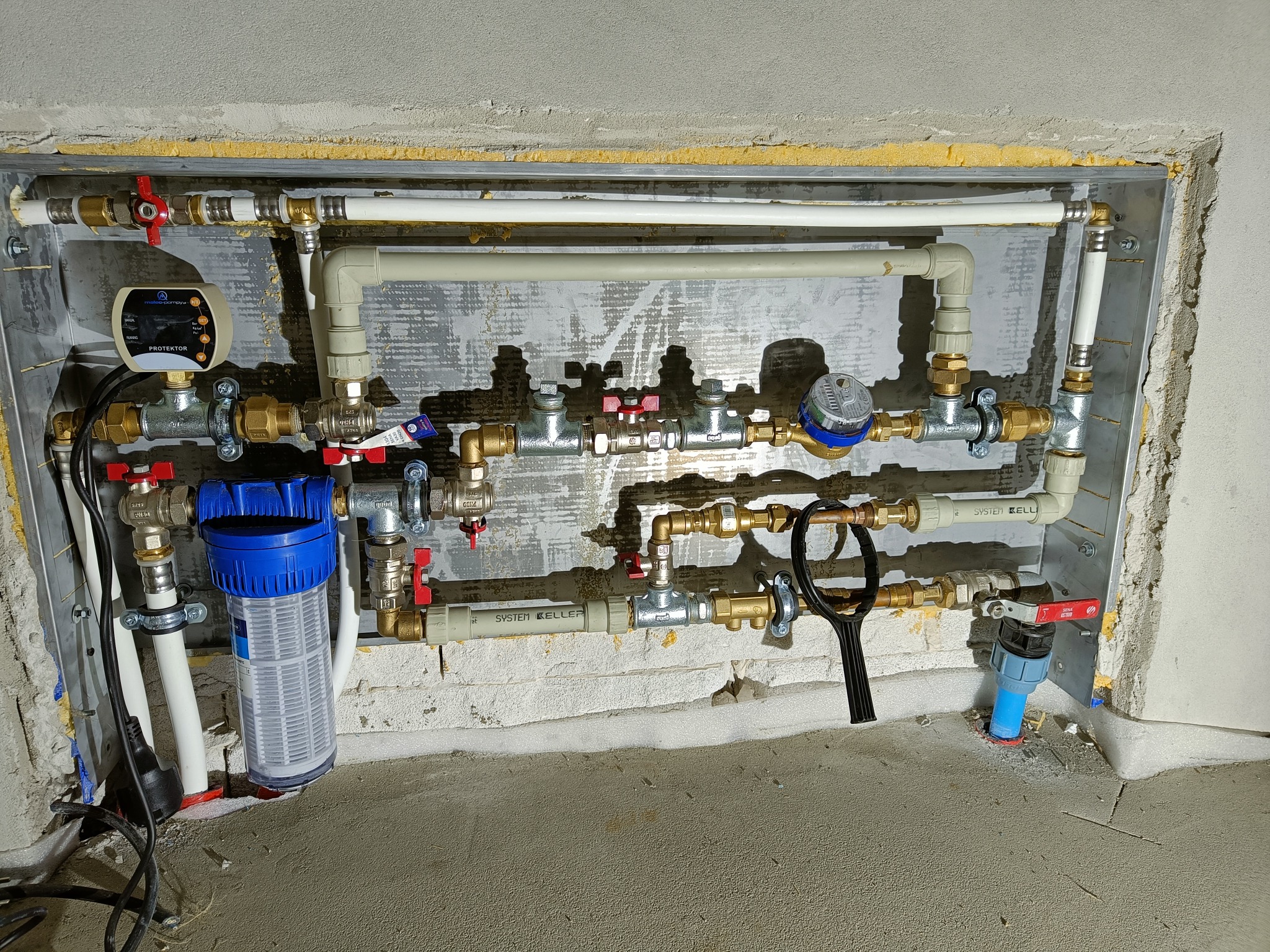 Złożona instalacja wodna wnęce ściennej, obejmująca rury z tworzywa i mosiądzu, zawory z czerwonymi uchwytami, filtr wody z niebieską obudową, wodomierz oraz urządzenie sterujące, widoczne ślady...