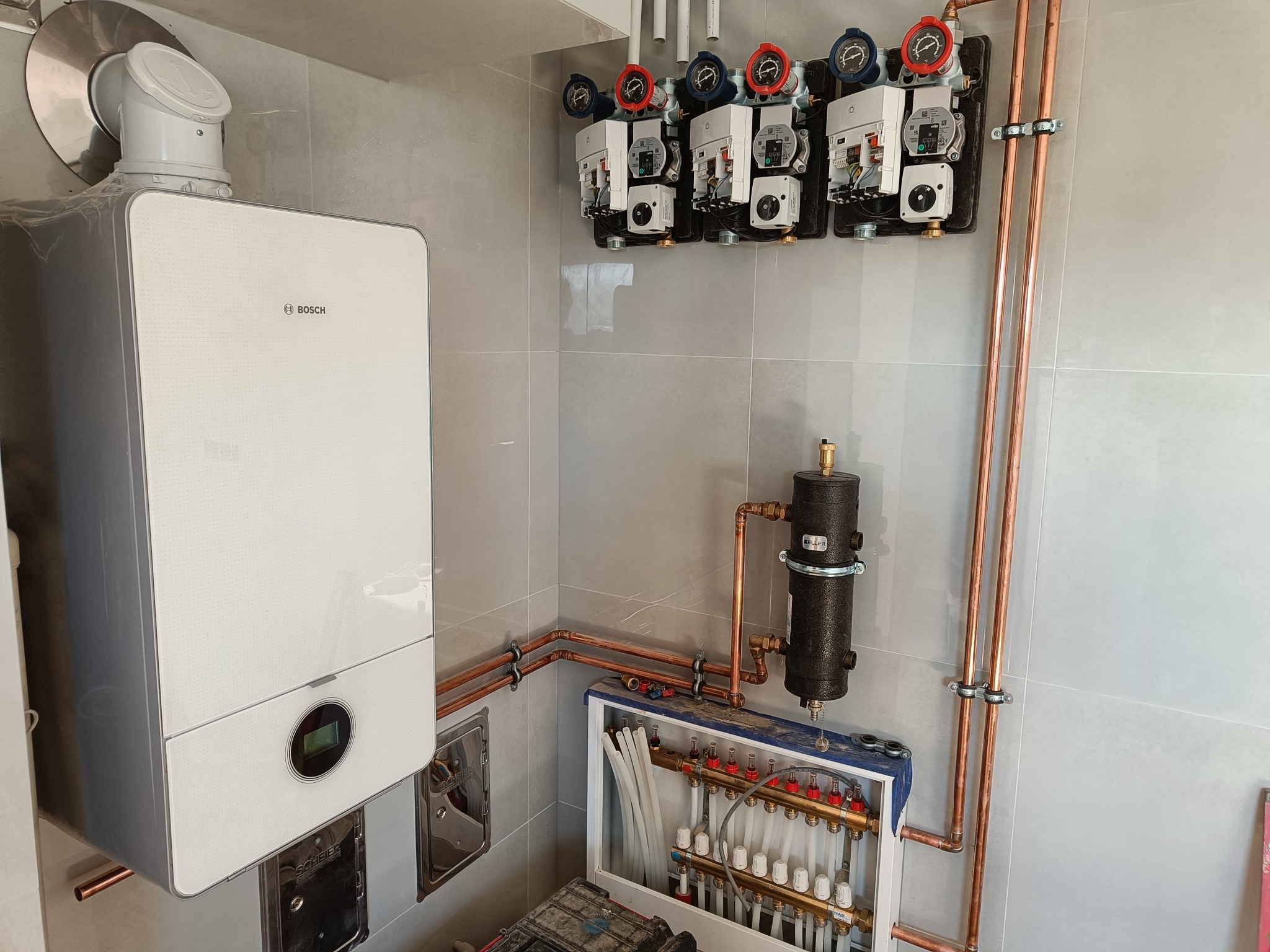 Nowoczesny piec gazowy Bosch z kompletną instalacją centralnego ogrzewania, obejmującą rozdzielacz podłogowy, pompy obiegowe i rury miedziane w nowym pomieszczeniu z szarymi płytkami.
