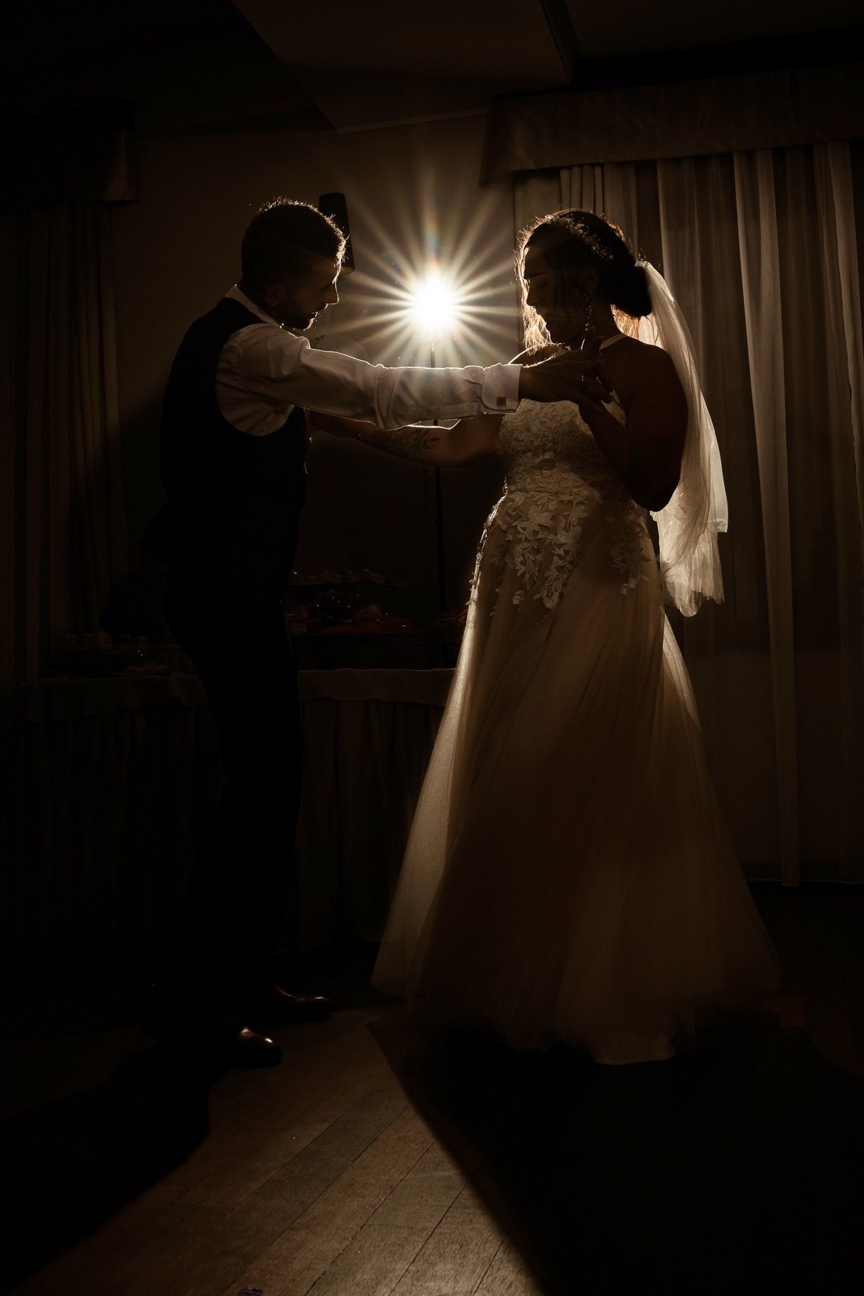 Para młoda w tańcu, silne oświetlenie z tyłu tworzy efektowne flary, panna młoda w sukni z koronkowymi aplikacjami i welonem, pan młody w kamizelce, ciemne tło z zasłonami.