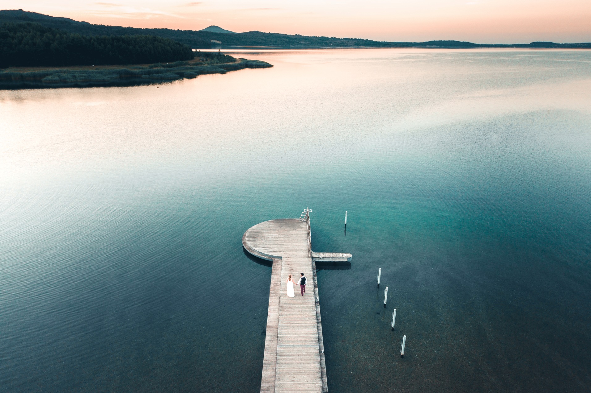 Zdjęcie z drona przedstawia parę młodą idącą po drewnianym molo w kierunku wody, zalesiony brzeg i góry w tle, oświetlone zachodzącym słońcem.