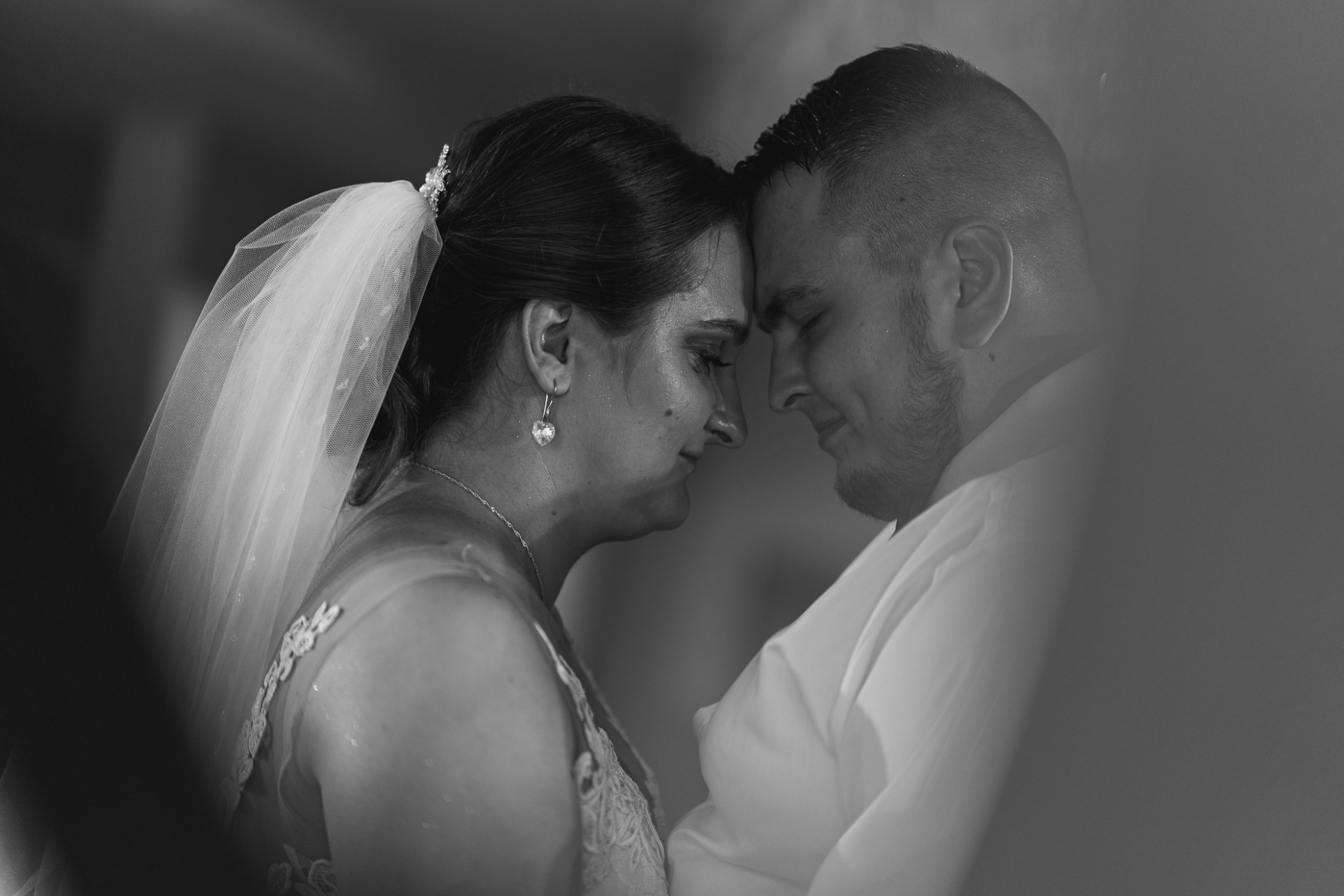 Czarno-białe zbliżenie pary młodej, czoła dotykają się, panna młoda w welonie i z kolczykami w kształcie serc, pan młody w białej koszuli.