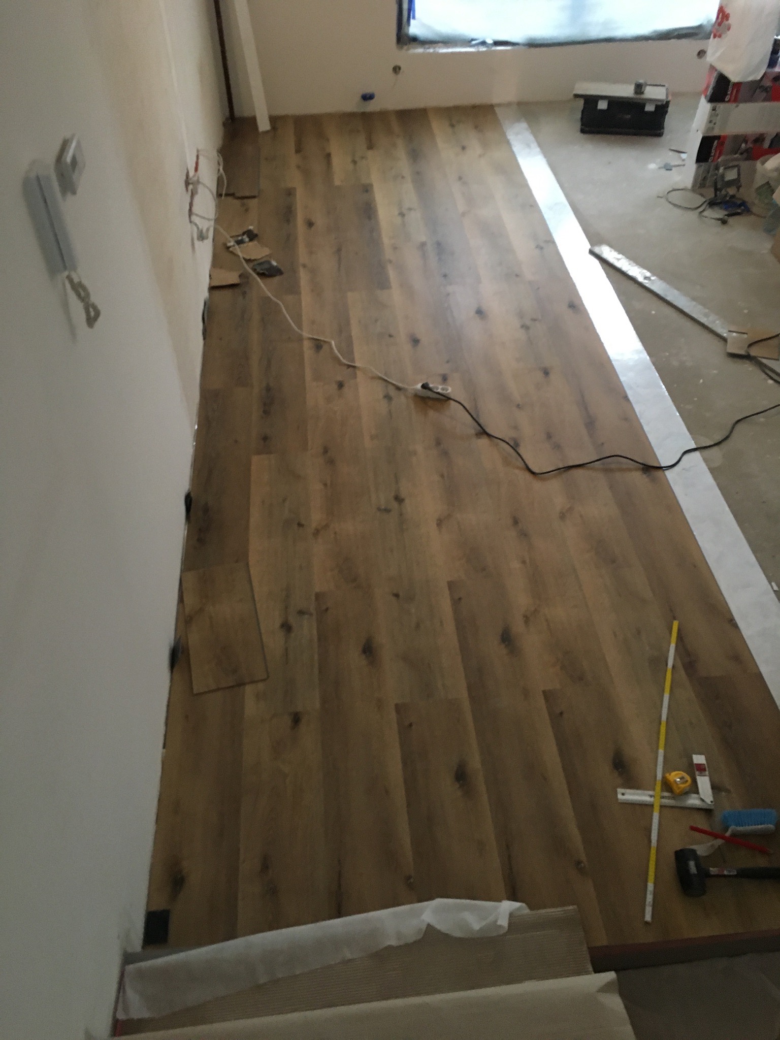 Układanie paneli podłogowych w trakcie prac, widoczne narzędzia: młotek, miarka, listwa pomiarowa, kabel zasilający, częściowo ułożona podłoga z paneli, biała ściana w tle.