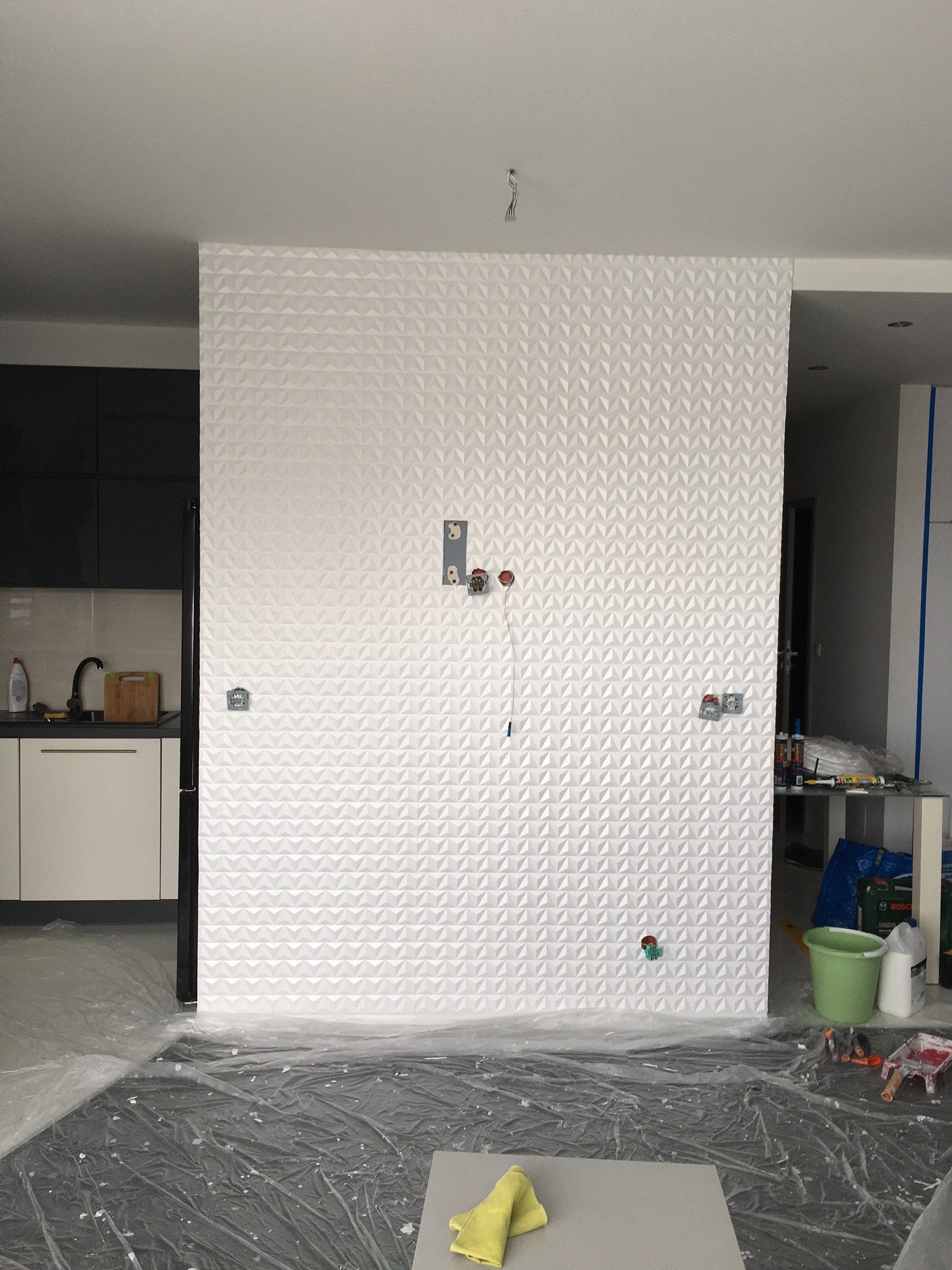 Ściana wykończona białą, trójwymiarową tapetą strukturalną z geometrycznym wzorem, zabezpieczona folią malarską na podłodze, widoczne puszki elektryczne i wystające przewody, w tle fragment kuchni...