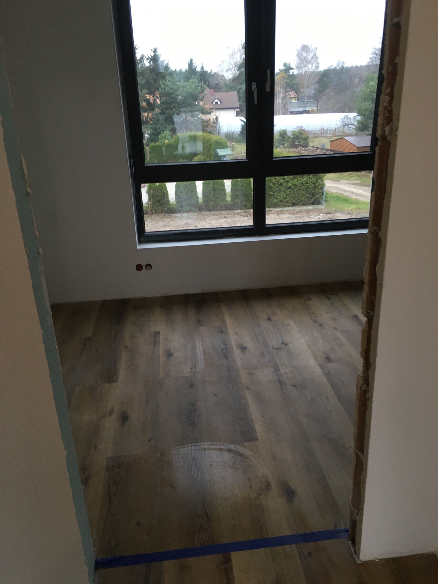 Nowo położona, brązowa podłoga panelowa w pustym pokoju z widokiem na ogród przez duże, czarne okno. Ściany w trakcie przygotowania do malowania, widoczne ślady po usuniętej taśmie malarskiej...