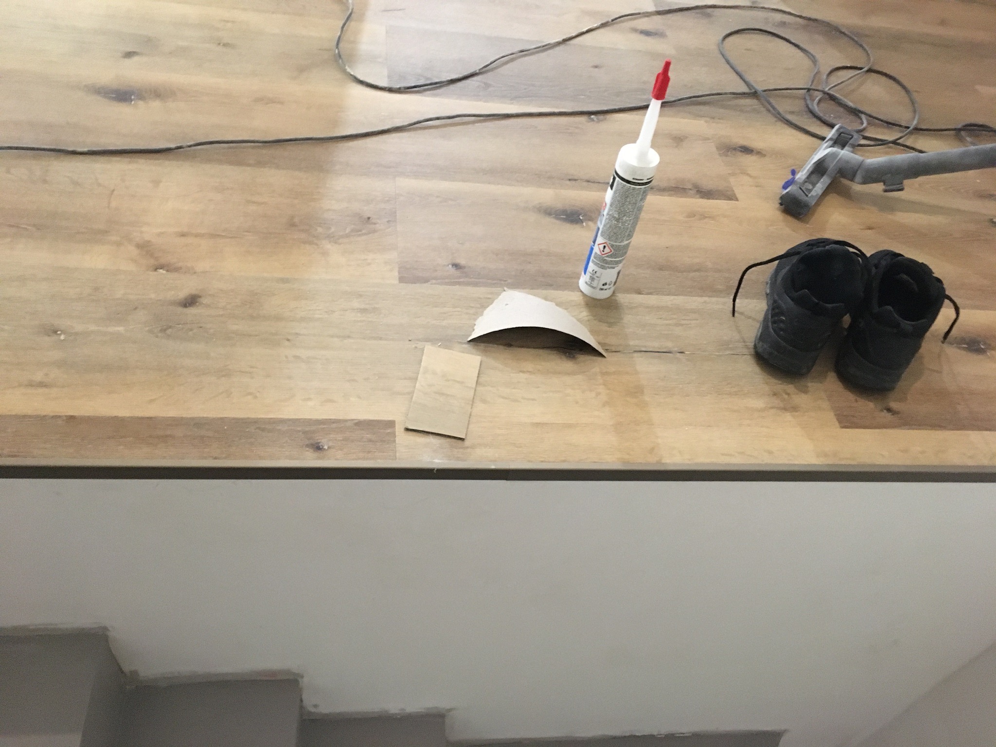 Układanie paneli podłogowych: widoczny fragment podłogi, tuba z uszczelniaczem, para butów roboczych, odkurzacz i kabel elektryczny.
