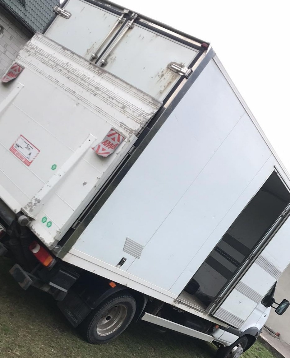 Biały samochód dostawczy z otwartymi tylnymi drzwiami, zaparkowany na trawie, widoczne ślady użytkowania na karoserii.