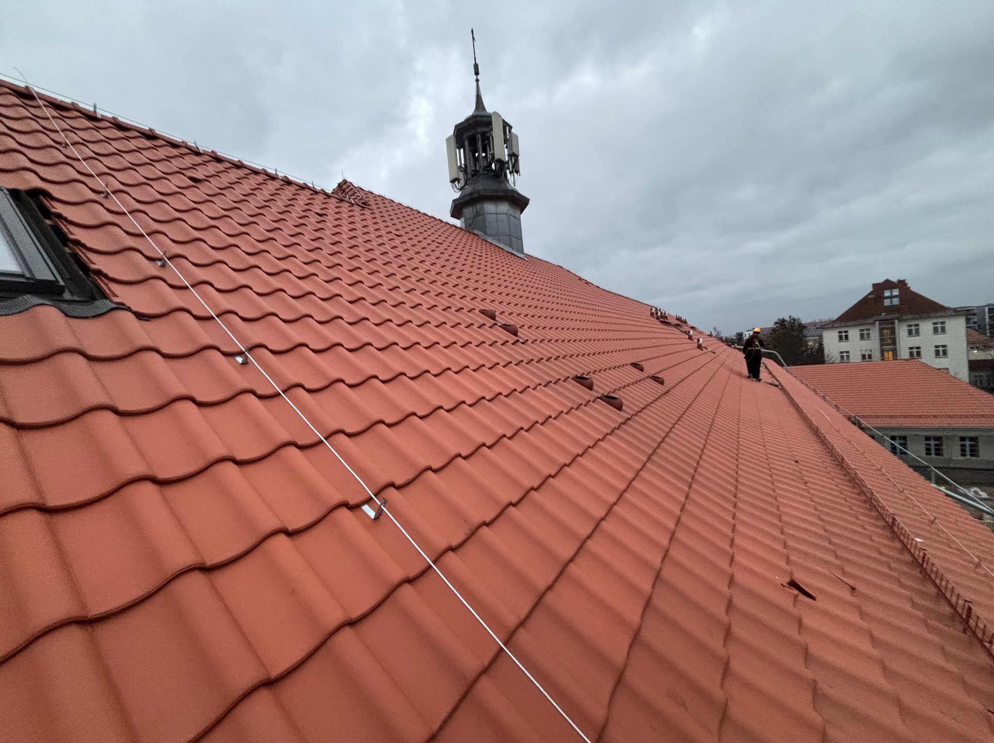 Czerwona dachówka ceramiczna na dachu budynku z wieżą w tle, zabezpieczona linką. Widoczne okno dachowe i fragment sylwetki osoby w oddali. Pochmurne niebo.